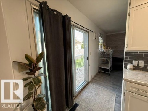 Sliding glass door leading to an outdoor deck, complemented by wood-finish flooring and a kitchen area featuring white cabinetry, a subway tile backsplash, and a light-toned countertop - 4917 45 Avenue, Vegreville, AB - Indoor Photo Showing Other Room