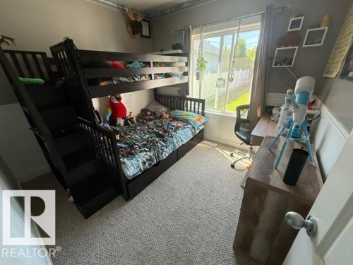 Carpeted room featuring a window with natural light, a dark wood-finish bunk bed with built-in storage drawers, and a light wood-finish desk with a desk chair - 4917 45 Avenue, Vegreville, AB - Indoor Photo Showing Bedroom