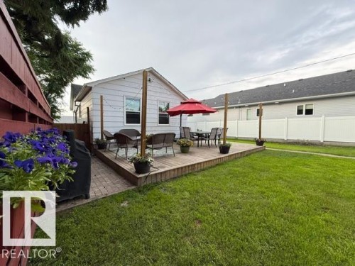 Rear exterior featuring an elevated wood deck, a patio area with paver stones, white vinyl siding, and a manicured lawn - 4917 45 Avenue, Vegreville, AB - Outdoor With Deck Patio Veranda