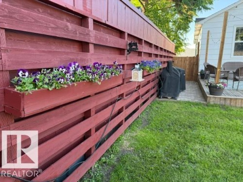 Red-stained wood privacy fence with integrated flower boxes featuring vibrant pansies, beside a paver patio and wood deck - 4917 45 Avenue, Vegreville, AB - Outdoor With Deck Patio Veranda