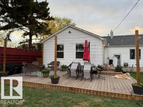 Spacious rear wood-finish deck with integrated steps, featuring a red privacy fence, surrounding green lawn, and exterior wall lighting - 4917 45 Avenue, Vegreville, AB - Outdoor With Deck Patio Veranda