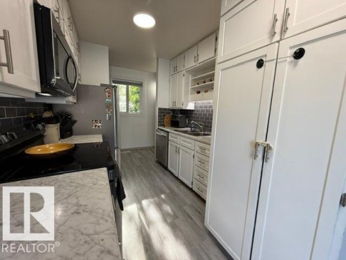 Galley kitchen featuring white cabinetry with brushed nickel hardware, light wood-finish flooring, and a dark tile backsplash - 4917 45 Avenue, Vegreville, AB - Indoor Photo Showing Kitchen