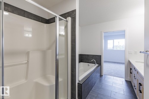 Bathroom featuring a glass-enclosed shower, a built-in soaking tub with dark tile surround, and a double vanity with light wood cabinetry and white countertops - 6308 65 Avenue, Beaumont, AB 