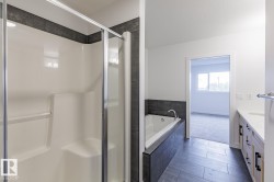 Bathroom featuring a glass-enclosed shower, a built-in soaking tub with dark tile surround, and a double vanity with light wood cabinetry and white countertops - 