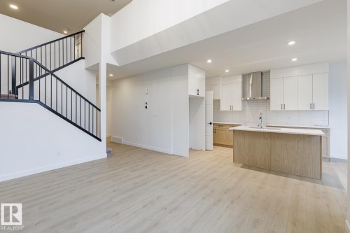 Open-concept kitchen and living area featuring light wood-finish flooring, a contemporary staircase with black metal railings, recessed lighting, white perimeter cabinetry, and an island with a stainless steel sink and a light-toned countertop - 6308 65 Avenue, Beaumont, AB 