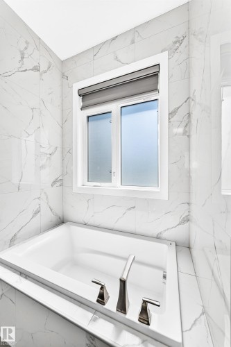 Soaking tub with brushed nickel fixtures, surrounded by large format tile with marble-look veining, and a window featuring a roller shade - 1781 Melrose Crescent, Edmonton, AB 