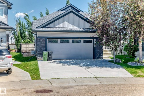 Contemporary residential exterior featuring a light gray garage door with upper window inserts, dark stone veneer accents, light gray horizontal siding, and a dark shingled roof with a prominent gable - 1781 Melrose Crescent, Edmonton, AB 
