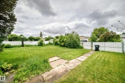 Expansive lawn area featuring a concrete paver pathway, mature perimeter trees, and a combination of white picket and chain-link fencing - 