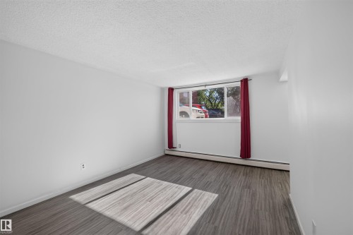 Spacious room featuring wood-finish flooring, white walls, and a large window with red drapes - 6 10230 122 Street Nw, Edmonton, AB 