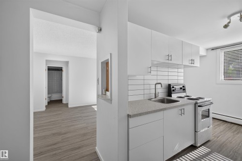 Compact kitchen featuring white cabinetry with brushed nickel hardware, a stone-finish countertop, stainless steel sink with a gooseneck faucet, and a subway tile backsplash - 6 10230 122 Street Nw, Edmonton, AB 