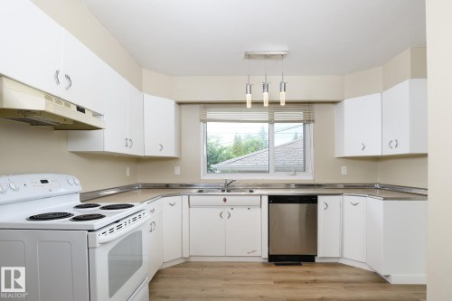 Well-appointed kitchen featuring white cabinetry, light wood-finish flooring, a stainless steel dishwasher, a white electric range, and a dual-basin sink with a window overlooking exterior greenery - 9607 75 Street, Edmonton, AB 