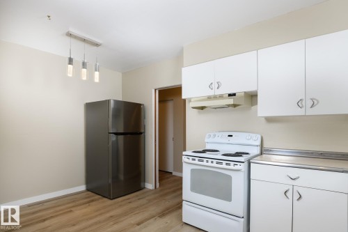 Kitchen space featuring wood-finish flooring, a stainless steel refrigerator, white cabinetry with silver hardware, a white electric range, and a brushed nickel linear pendant light fixture - 9607 75 Street, Edmonton, AB 