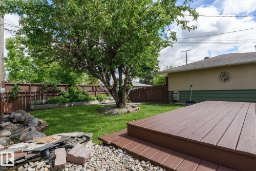 Expansive backyard featuring a large mature tree, a brown privacy fence with lattice accents, a multi-level wood-finish deck, and a rock garden with varied stone elements - 9607 75 Street, Edmonton, AB 
