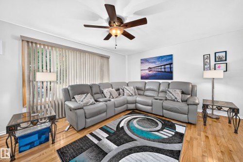 Light-filled living area featuring wood-finish flooring, a ceiling fan with integrated lighting, and a large window with vertical blinds - 11708 138 Avenue Nw, Edmonton, AB 