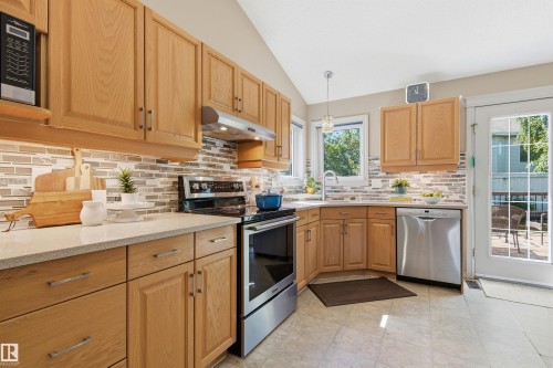 Bright kitchen featuring light wood cabinetry, stone-finish countertops, stainless steel appliances, a mosaic tile backsplash, and a glass-paned exterior door - 11715 11B Avenue, Edmonton, AB 