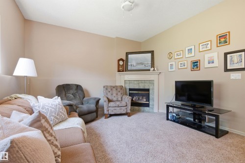 Living room featuring a fireplace with a white mantel and green tile surround, neutral carpeting, and sloped ceilings - 11715 11B Avenue, Edmonton, AB 