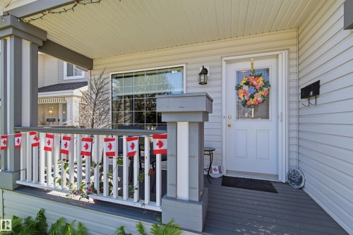Covered front porch with grey decking and white railing, featuring light-colored siding, an exterior wall lantern, and a front entry door with a window insert - 11715 11B Avenue, Edmonton, AB 