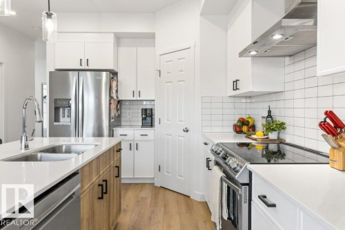 Modern kitchen featuring a large island with an undermount double-basin sink, stainless steel appliances, white flat-panel cabinetry with black hardware, white subway tile backsplash, and wood-finish flooring - 25 Nettle Crescent, St. Albert, AB 