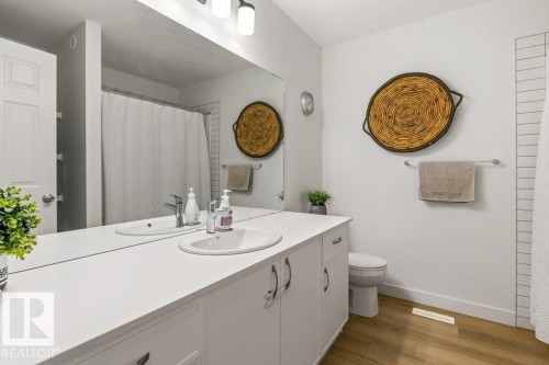 Bathroom featuring a dual-sink vanity with white cabinetry, a large wall-mounted mirror, wood-finish flooring, and a white toilet - 25 Nettle Crescent, St. Albert, AB 