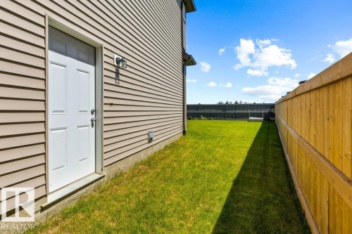 Side yard featuring a white entry door with overhead transom, tan horizontal siding, and a wooden privacy fence enclosing a grass lawn - 25 Nettle Crescent, St. Albert, AB 