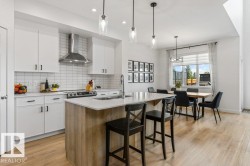 Open-concept kitchen featuring white cabinetry, a subway tile backsplash, stainless steel range hood, and an island with a light-toned countertop - 