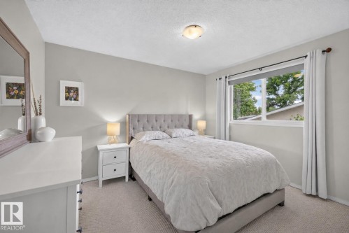 Carpeted bedroom featuring a window with roller shade and drapes, a tufted headboard, and a white nightstand with a built-in lamp - 244 Knottwood Road N, Edmonton, AB 