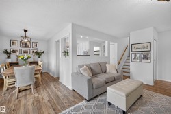 Open-concept living area featuring wood-finish flooring, white wall finishes, a prominent staircase with a wooden handrail, and a pass-through window to an additional room - 