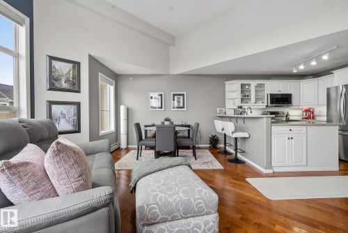 Open-concept living area featuring high vaulted ceilings, large windows, wood-finish flooring, a dining space, and a kitchen with white cabinetry and stainless steel appliances - 421 9730 174 Street, Edmonton, AB 
