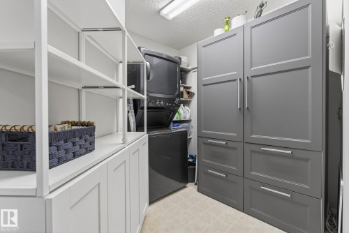 Laundry room with a stacked washer and dryer, white open shelving, white lower cabinetry, and gray closed cabinetry with drawers - 421 9730 174 Street, Edmonton, AB 