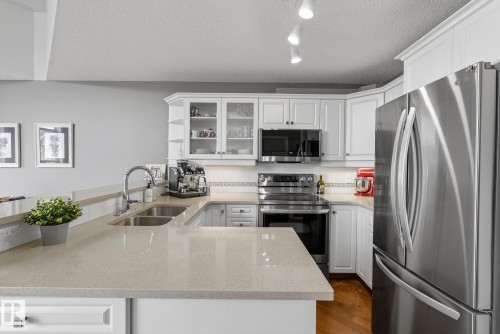 U-shaped kitchen featuring stone countertops, stainless steel appliances, white cabinetry with glass-front uppers, and a double basin sink with a gooseneck faucet - 421 9730 174 Street, Edmonton, AB 