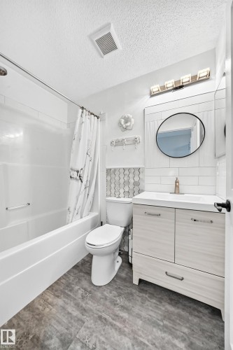 White bathroom featuring a single vanity with light wood-finish cabinetry, a white countertop, and a round black-framed mirror - 146 5604 199 Street, Edmonton, AB 