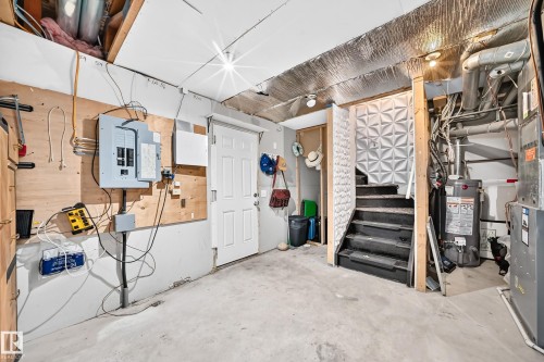 Utility area featuring a concrete floor, exposed ceiling joists, electrical panel, water heater, and an adjacent staircase with textured paneling - 146 5604 199 Street, Edmonton, AB 