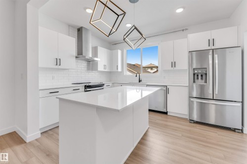 Modern kitchen featuring a central island with a light-toned countertop, white cabinetry with dark hardware, stainless steel appliances, a subway tile backsplash, and wood-finish flooring - 1208 14 Ave Nw, Edmonton, AB 