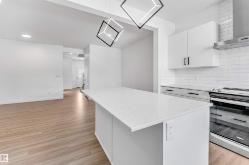 Open-concept kitchen featuring a spacious island with a light-toned countertop, white cabinetry with black hardware, a stainless steel range hood, and subway tile backsplash - 1208 14 Ave Nw, Edmonton, AB 