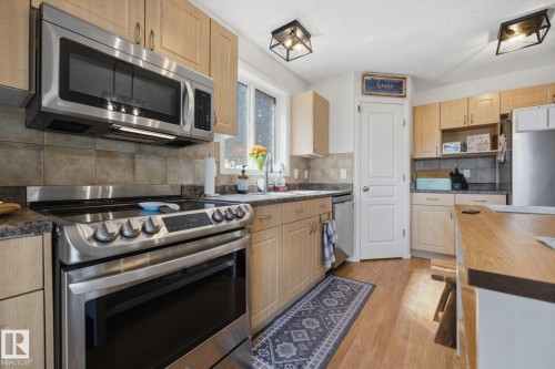 Kitchen featuring light wood cabinetry, stainless steel appliances, a tile backsplash, dark countertops, and wood-finish flooring - 60 Leyland Way, Spruce Grove, AB 