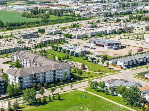Aerial perspective showcasing a multi-story residential building with balconies, alongside modern townhomes, commercial properties, and a green space with a sports field - 60 Leyland Way, Spruce Grove, AB 
