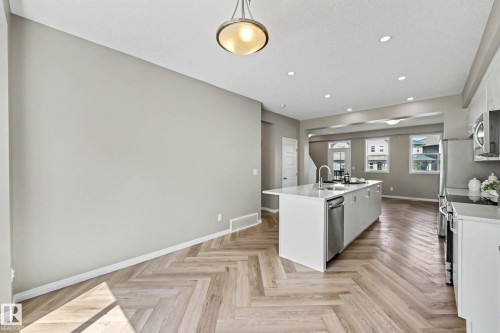 Open-concept living space featuring wood-finish flooring in a herringbone pattern, a central kitchen island with an undermount sink, recessed lighting, and stainless steel appliances - 53 2072 Wonnacott Way, Edmonton, AB 