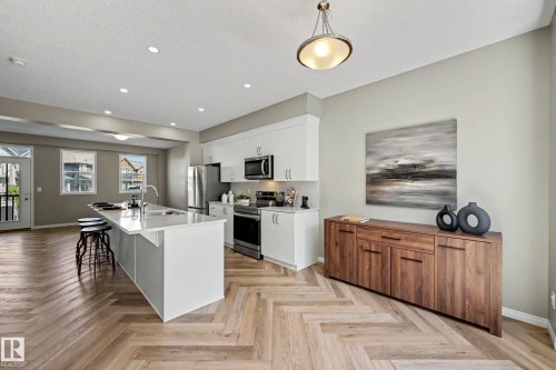 Open-concept kitchen featuring an island with a breakfast bar, white cabinetry, stainless steel appliances, and wood-finish flooring in a herringbone pattern - 53 2072 Wonnacott Way, Edmonton, AB 