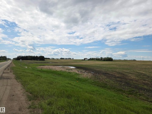 Expansive agricultural land featuring a paved road, green grass roadside, a visible body of water, and distant structures - 24209 Hwy 625, Beaumont, AB 
