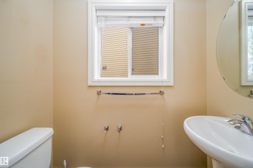Compact bathroom featuring a white porcelain toilet, a pedestal sink with a chrome faucet, a round wall-mounted mirror, and a window with white trim - 46 15151 43 Street, Edmonton, AB 