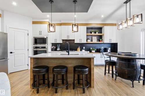 Contemporary kitchen featuring a large central island with seating, integrated stainless steel appliances, a dark backsplash, white cabinetry with contrasting wood accents, and light wood-finish flooring - 7 Tenor Link, Spruce Grove, AB 