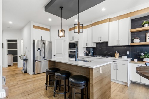 Modern kitchen featuring a central island with countertop seating, stainless steel appliances, light wood-finish flooring, white cabinetry, and a dark tile backsplash - 7 Tenor Link, Spruce Grove, AB 