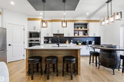 Contemporary kitchen featuring a large central island with seating, integrated stainless steel appliances, a dark backsplash, white cabinetry with contrasting wood accents, and light wood-finish flooring - 