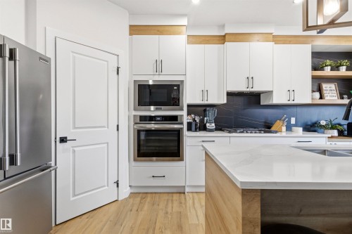Modern kitchen featuring white cabinetry, light wood-finish flooring, a stainless steel refrigerator, built-in microwave and oven, and a kitchen island with a light-toned countertop - 7 Tenor Link, Spruce Grove, AB 