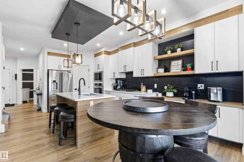 Modern kitchen featuring white cabinetry, light wood-finish flooring, a central island with bar seating, integrated stainless steel appliances, and a black stone-finish backsplash - 7 Tenor Link, Spruce Grove, AB 