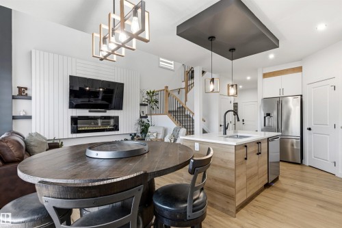 Open-concept living space featuring wood-finish flooring, a contemporary kitchen island with a white countertop and dark fixtures, an electric fireplace with a white shiplap-style surround, stainless steel appliances, and a modern rectangular chandelier - 7 Tenor Link, Spruce Grove, AB 
