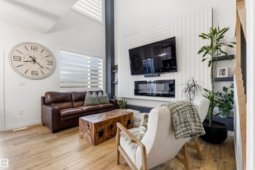Vaulted ceilings enhance this living area featuring wood-finish flooring, a wall-mounted electric fireplace, and a large window with horizontal blinds - 7 Tenor Link, Spruce Grove, AB 