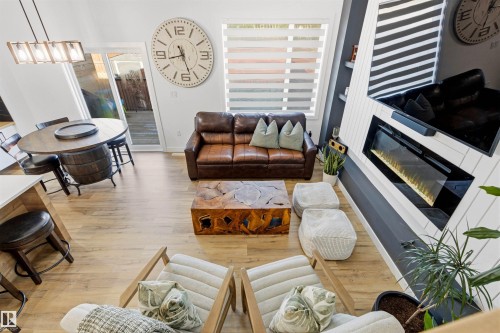 Open-concept living space featuring wood-finish flooring, a contemporary fireplace with a sleek surround, an attached dining area with a unique barrel-base table, and large window with horizontal blinds - 7 Tenor Link, Spruce Grove, AB 