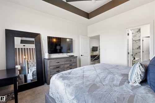 Bedroom featuring a recessed tray ceiling with accent paint, carpeted flooring, and a door opening to a bathroom with marble-patterned wall tile - 7 Tenor Link, Spruce Grove, AB 