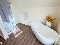 Freestanding oval soaking tub with chrome fixtures, curved glass shower enclosure, wood-finish flooring, and crisp white baseboards - 